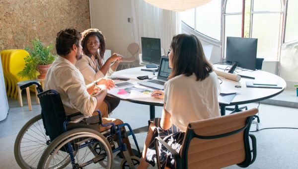 3 individuals sitting at board room table discussing a business plan one male in a wheelchair